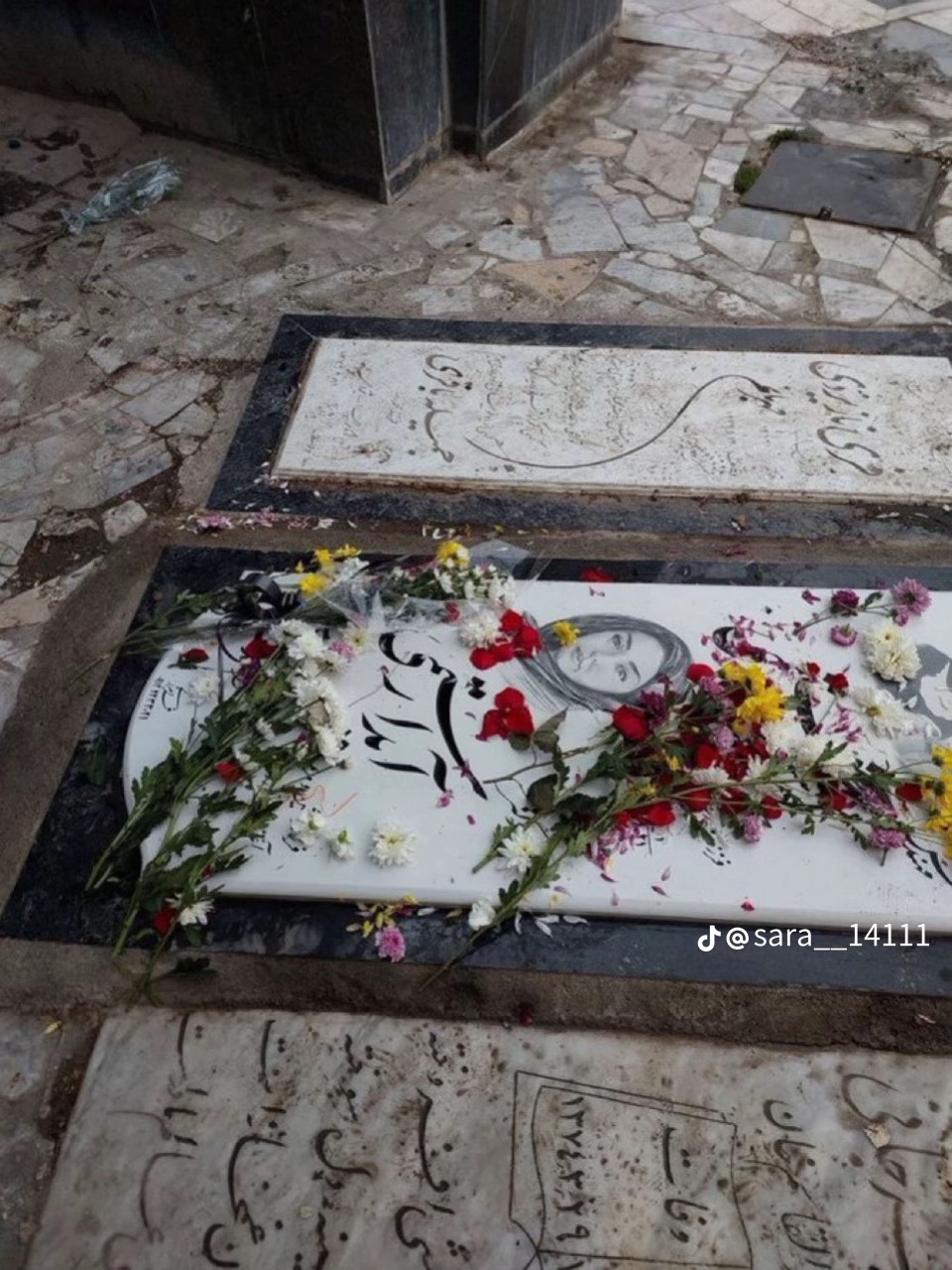 La tombe d’Aida Rostami à Téhéran, dalle de marbre blanc avec son portrait gravé, jonchée de roses rouges et de marguerites.